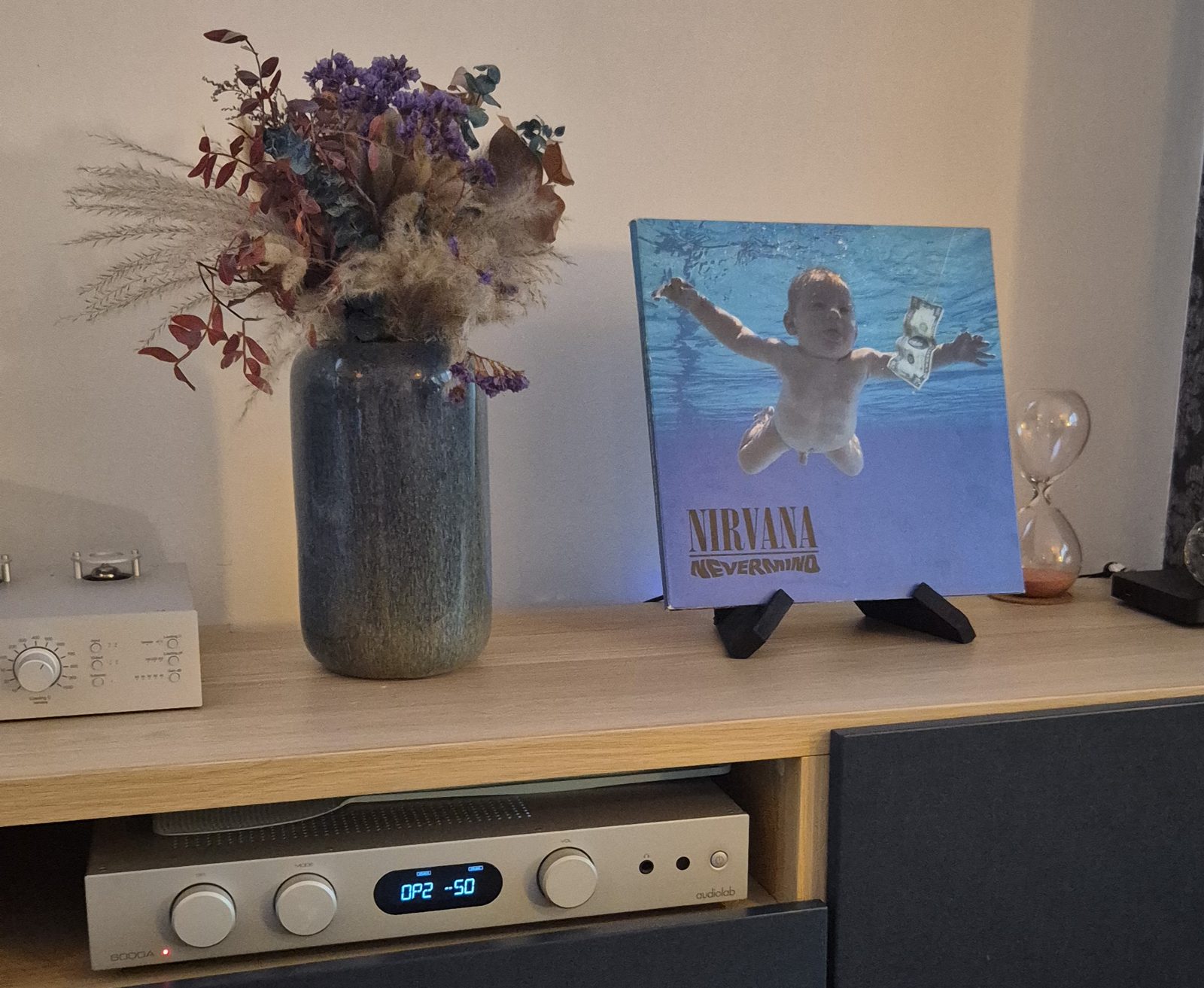 The Nevermind LP standing on a small black stand on a wooden sideboard, next to a vase of dried flowers, with an amplifier in the shelf below